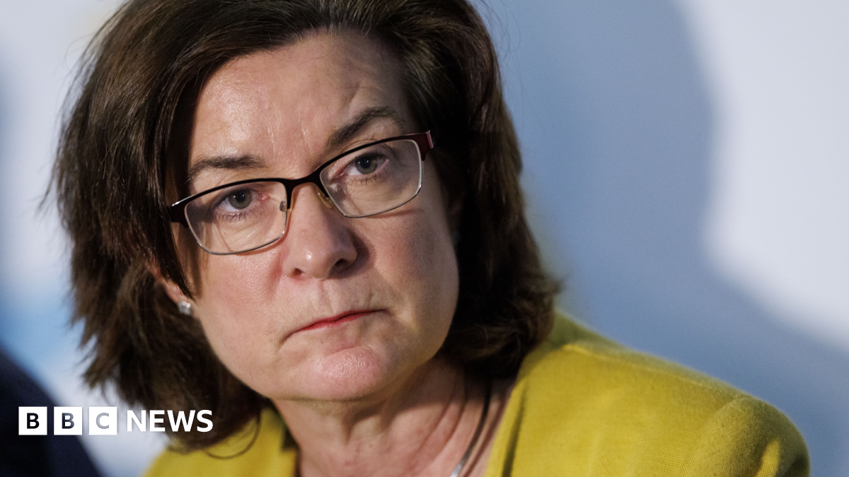 First Minister Eluned Morgan is looking past the camera and her head turned, wearing a yellow jacket and glasses.