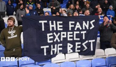 Fans hold a bedsheet banner saying 'The Fans Expect The Best' in a football stadium.