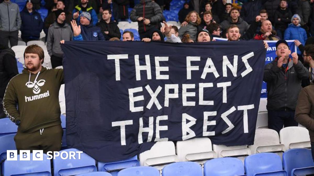 Fans hold a bedsheet banner saying 'The Fans Expect The Best' in a football stadium.