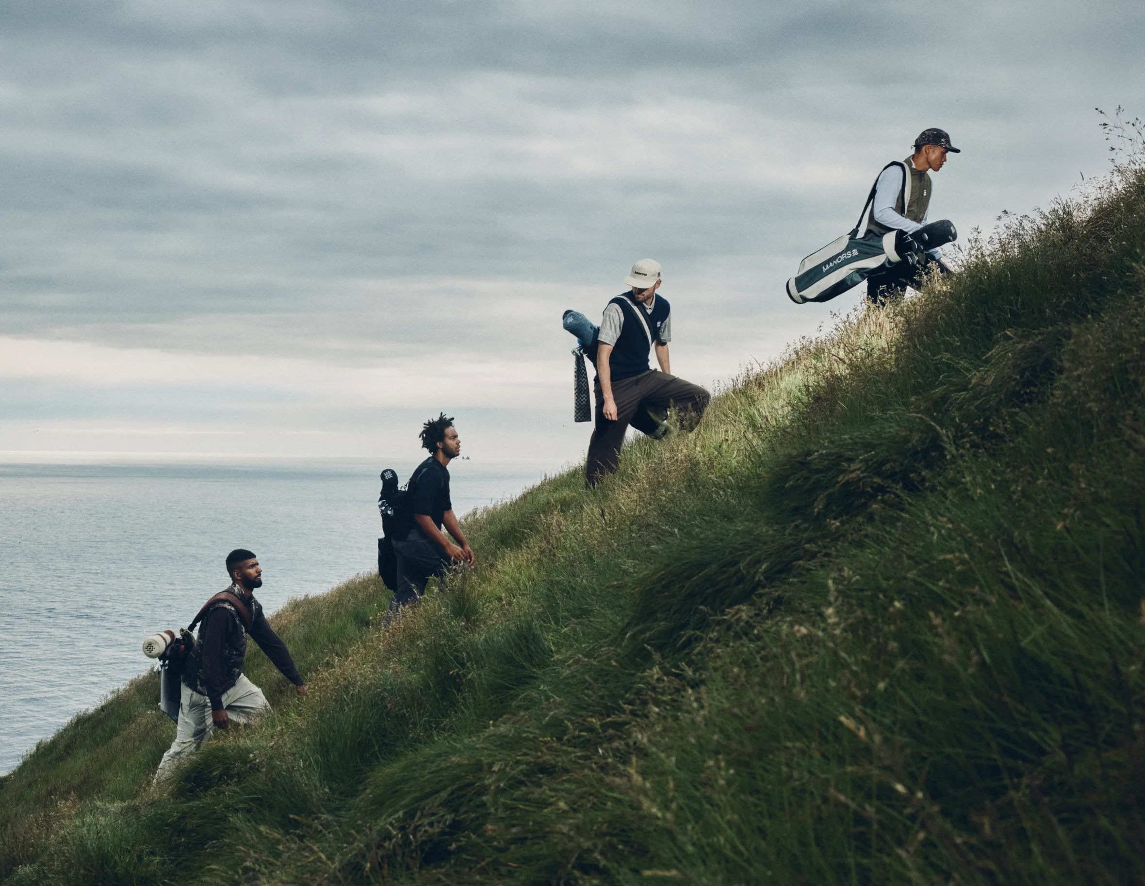 Four men carrying golf bags climb a grassy hill overlooking the ocean.