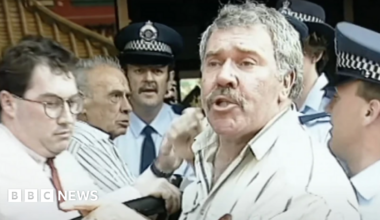Still from the video, showing Jack Karlson, wearing a beige shirt, surrounded by police and men in shirts trying to hussle him along as he speaks to the camera.