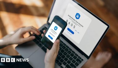 A person holds a smartphone over a laptop to complete a two-factor authentication process. Both screens display security prompts and matching verification codes.