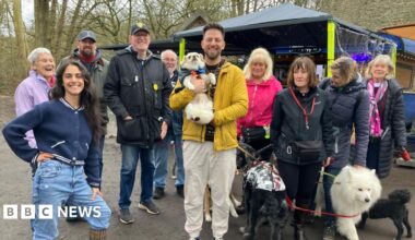 A group of people stand outside, in a park with their dogs. The coffee stand is in the background, with an awning for people to sit underneath.