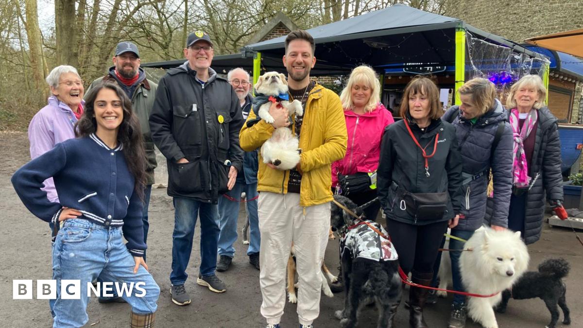 A group of people stand outside, in a park with their dogs. The coffee stand is in the background, with an awning for people to sit underneath.
