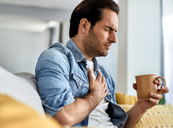 Man in denim shirt clutching chest in pain while sitting on couch holding a cup, highlighting diseases people should fear more.