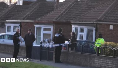 Five police officers stand guard in front of a property which has been cordoned off. The house is fronted by a metal rail and brick wall which has blue and white police tape attached to it. A row of other houses are visible in the background.