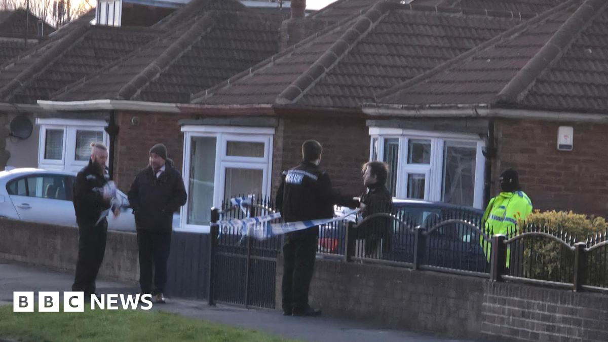 Five police officers stand guard in front of a property which has been cordoned off. The house is fronted by a metal rail and brick wall which has blue and white police tape attached to it. A row of other houses are visible in the background.