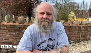 A man in his 60s sits with his arms folded and resting on a table outside. He has a mostly bald head with wispy grey hair and a long beard. He is wearing light blue T-shirt with a motif on it and text that reads "Bridgwater, Somerset". Behind him is a brick wall, a large yellow house and trees.