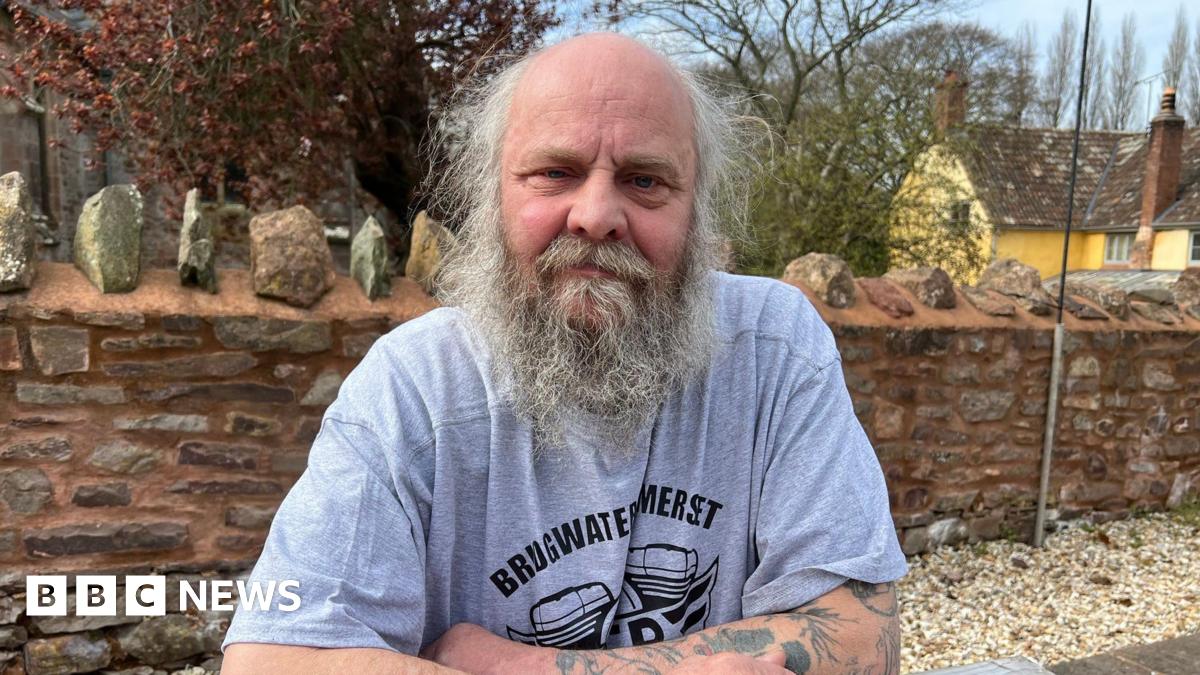 A man in his 60s sits with his arms folded and resting on a table outside. He has a mostly bald head with wispy grey hair and a long beard. He is wearing light blue T-shirt with a motif on it and text that reads "Bridgwater, Somerset". Behind him is a brick wall, a large yellow house and trees.