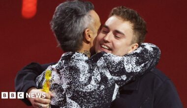 Robbie Williams has short grey hair and a black and white patterned formal jacket. He is embracing Sam Fender who has short brown hair and is holding a Brit Award. He is wearing a dark jacket. Olivia Dean is standing next to them with long dark hair and wearing a patterned black dress.
