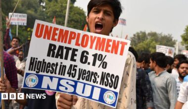 A young man wearing a white shirt holds a placard with slogans against unemployment and looks into the camera while shouting. Behind him are other protesters