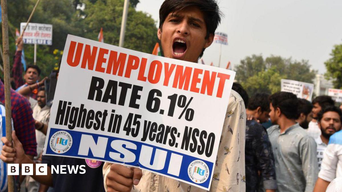 A young man wearing a white shirt holds a placard with slogans against unemployment and looks into the camera while shouting. Behind him are other protesters