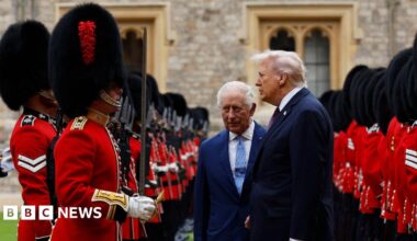 President Donald Trump and King Charles inspect a Guard of Honour together. The pair, both wearing dark suits, are stood between two rows of Beefeaters, dressed in red and with tall black headwear.