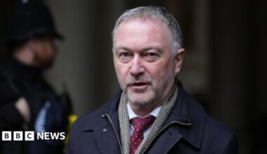 Communities secretary Steve Reed wearing a dark coloured coat, a check pattern scarf and a red tie. A police officer is stood in the background.