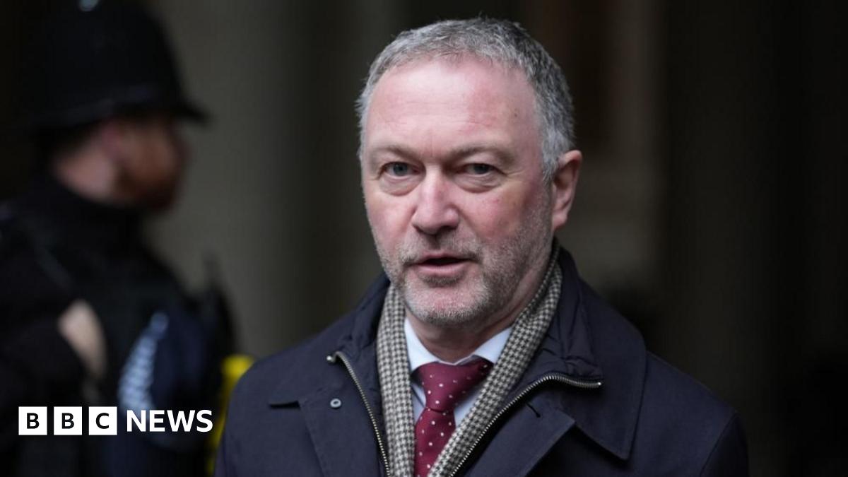 Communities secretary Steve Reed wearing a dark coloured coat, a check pattern scarf and a red tie. A police officer is stood in the background.
