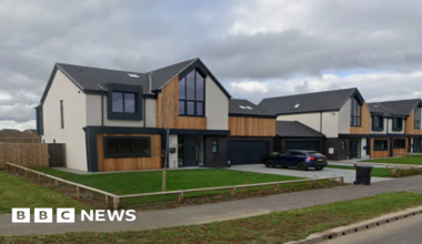 A row of modern detached homes featuring dark grey roofs, dark window frames, wood pannelling and grey and cream cladding. Each has a plain rectangle of grass at the front. The sky is cloudy.