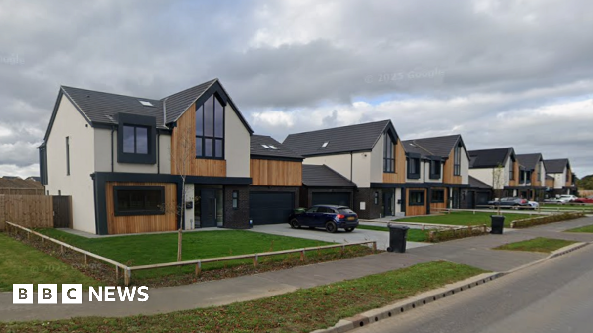 A row of modern detached homes featuring dark grey roofs, dark window frames, wood pannelling and grey and cream cladding. Each has a plain rectangle of grass at the front. The sky is cloudy.
