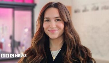 A woman with long black hair wearing a yellow knitted jumper looks at someone off-camera