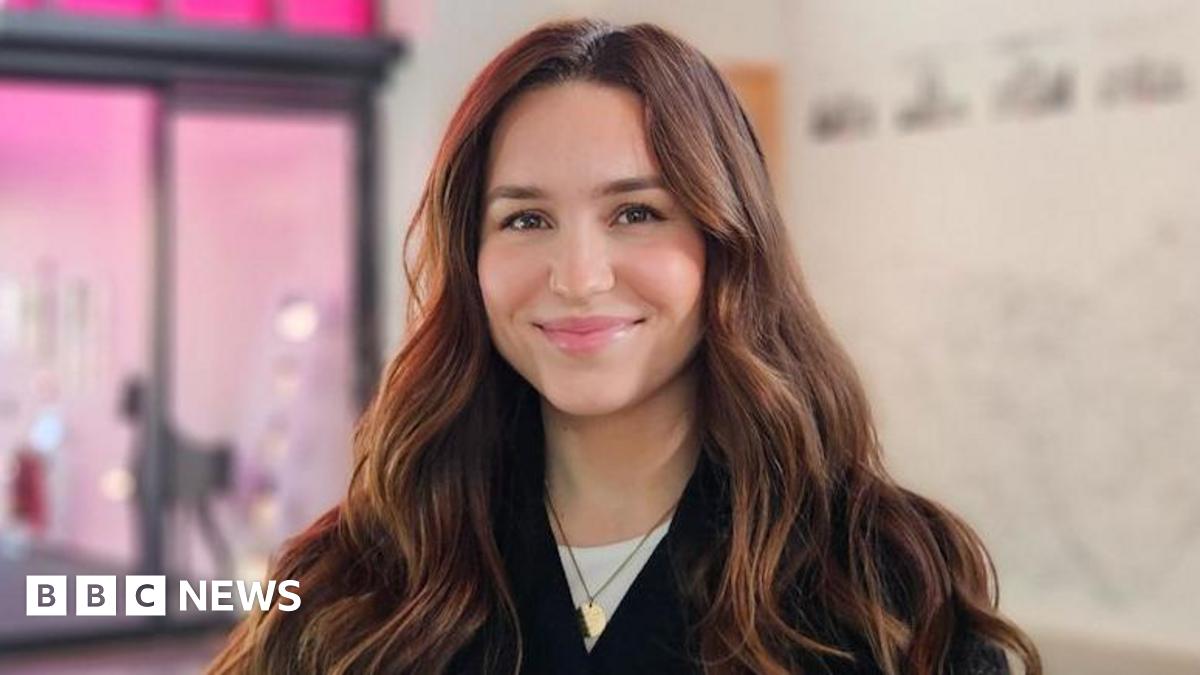 A woman with long black hair wearing a yellow knitted jumper looks at someone off-camera