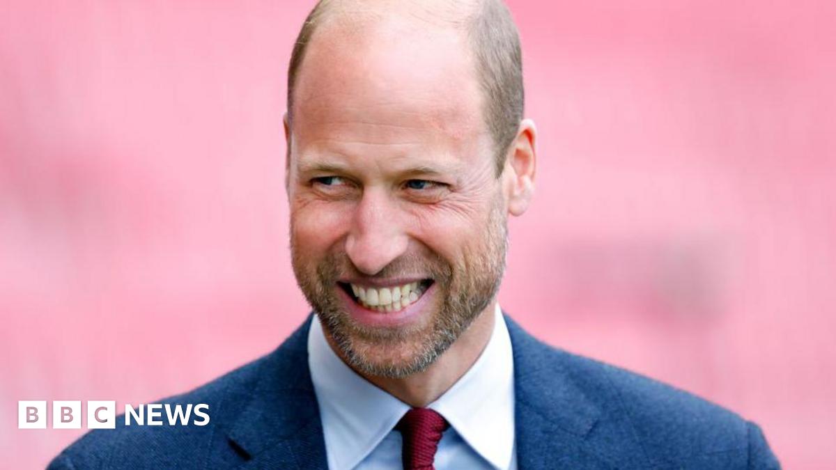 Prince William, wearing a navy suit and red tie, smiles