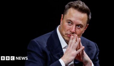 Elon Musk on a stage with his hands tented in front of his mouth as he listens to someone next to him.