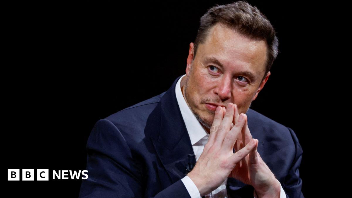 Elon Musk on a stage with his hands tented in front of his mouth as he listens to someone next to him.