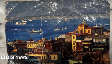 A dog-eared post card depicting a coastal community, with boats in a bay flanked by old, multi-storey homes.