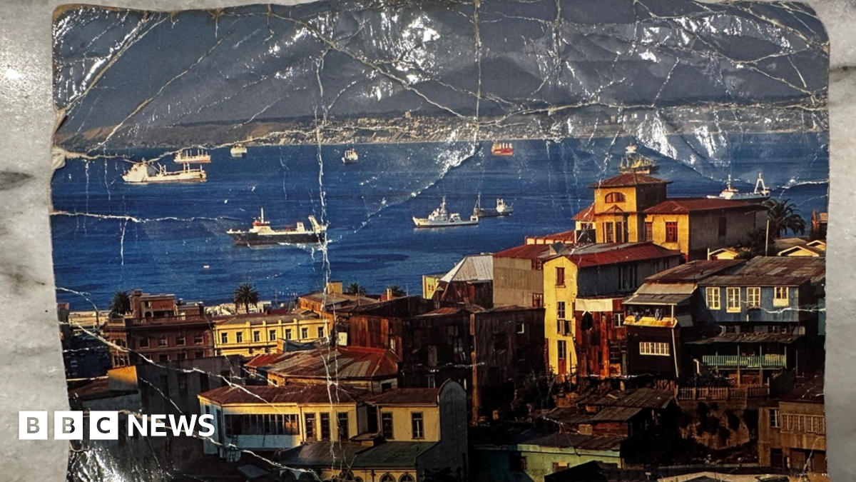 A dog-eared post card depicting a coastal community, with boats in a bay flanked by old, multi-storey homes.
