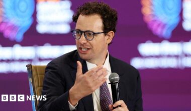 Dario Amodei, Anthropic's CEO, speaking on stake while holding a microphone, wearing a black suit jacket, white shirt and red and navy spotted neck tie.