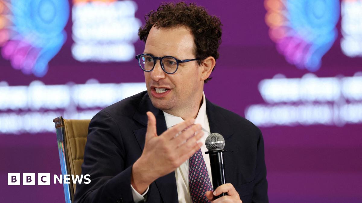 Dario Amodei, Anthropic's CEO, speaking on stake while holding a microphone, wearing a black suit jacket, white shirt and red and navy spotted neck tie.