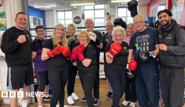 A group of elderly men and women dressed in dark gym gear hold up their boxing gloves, smiling.