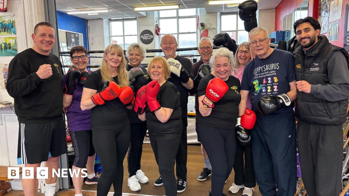 A group of elderly men and women dressed in dark gym gear hold up their boxing gloves, smiling.