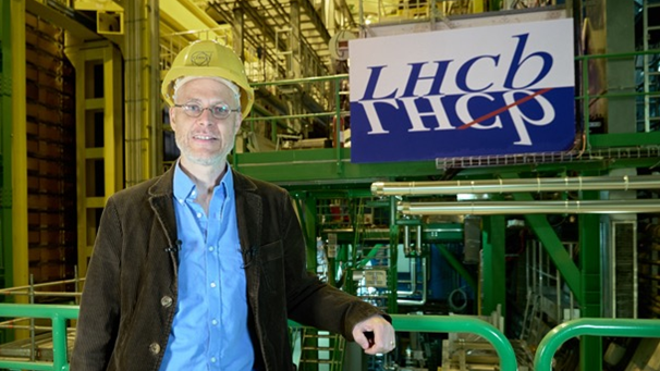 Prof Chris Parkes in a yellow hard hat standing in front of the LHCb experiment.