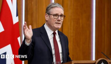 Sir Keir Starmer stands at a podium and raises his right hand. A large Union Jack is behind him.