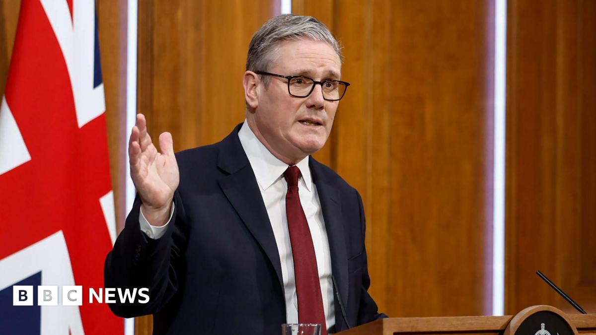 Sir Keir Starmer stands at a podium and raises his right hand. A large Union Jack is behind him.