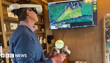 Ian Sexton is standing in a shed wearing virtual reality goggles while he looks at a screen which shows fields. One of the fields has a yellow grid in laid over it. In his hands are two handsets.