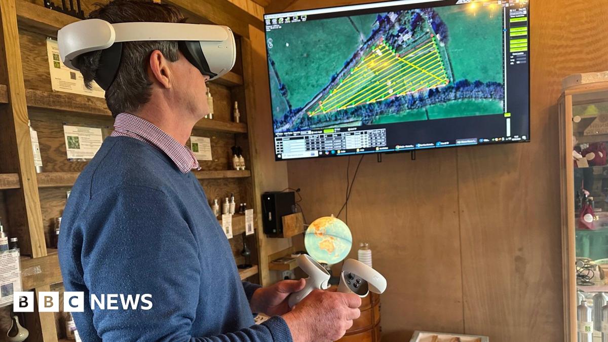 Ian Sexton is standing in a shed wearing virtual reality goggles while he looks at a screen which shows fields. One of the fields has a yellow grid in laid over it. In his hands are two handsets.