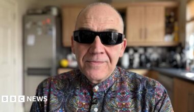 A man with balding short grey hair looks into the camera. He is wearing dark sunglasses and a multicoloured patterned shit. He is sitting in his kitchen.