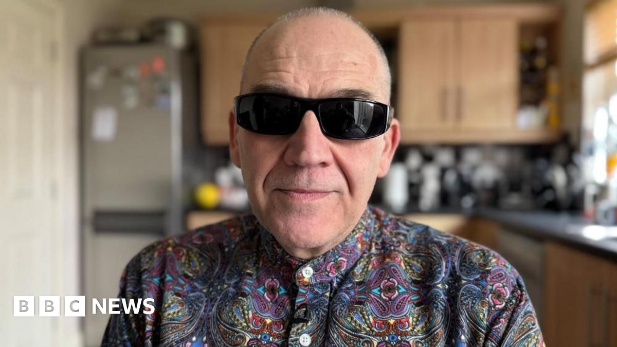 A man with balding short grey hair looks into the camera. He is wearing dark sunglasses and a multicoloured patterned shit. He is sitting in his kitchen.