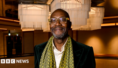 A man with short dark hair, a white shirt and dark jacket with a patterned yellow and black scarf around his neck and brown walls behind him