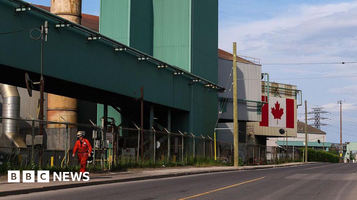 An image of a steel plant in Hamilton, Ontario. A sole worker in a red jumpsuit and a helmet is seen walking outside. The plant building is green, white and beige, and features a large Canadian flag on one panel.