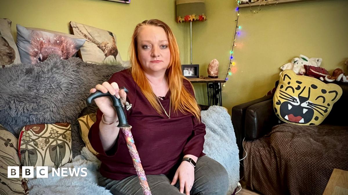 A woman in her 40s is sitting on a sofa in her living room holding a walking stick. She has long orange hair. Behind her is cushions and lighting.