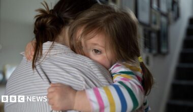 A woman with brown hair tied up and wearing a grey and white striped shirt hugs a young girl who is wearing a colourfully striped long-sleeved top and has her brown hair tied in pig tails