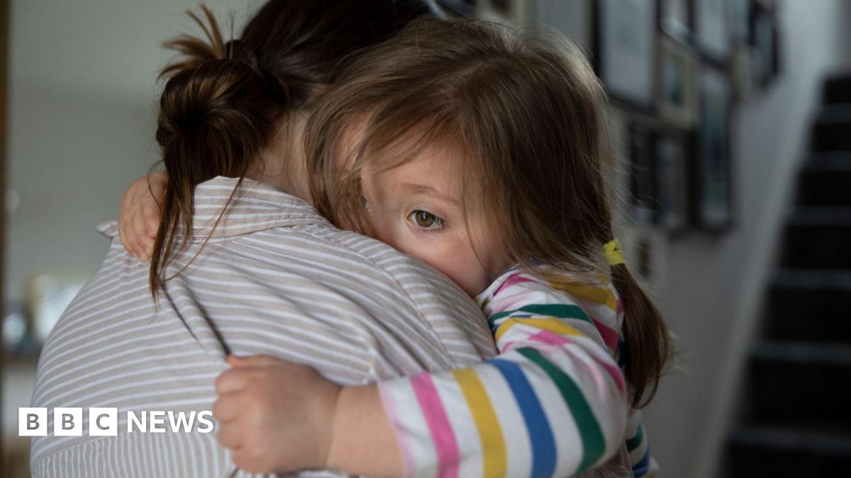 A woman with brown hair tied up and wearing a grey and white striped shirt hugs a young girl who is wearing a colourfully striped long-sleeved top and has her brown hair tied in pig tails