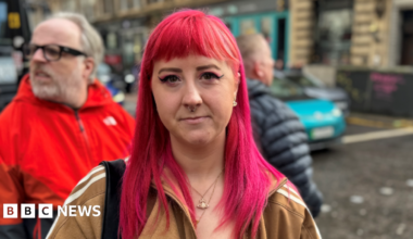 A woman with pink hair stands in the middle of the street with a light brown Adidas coat on