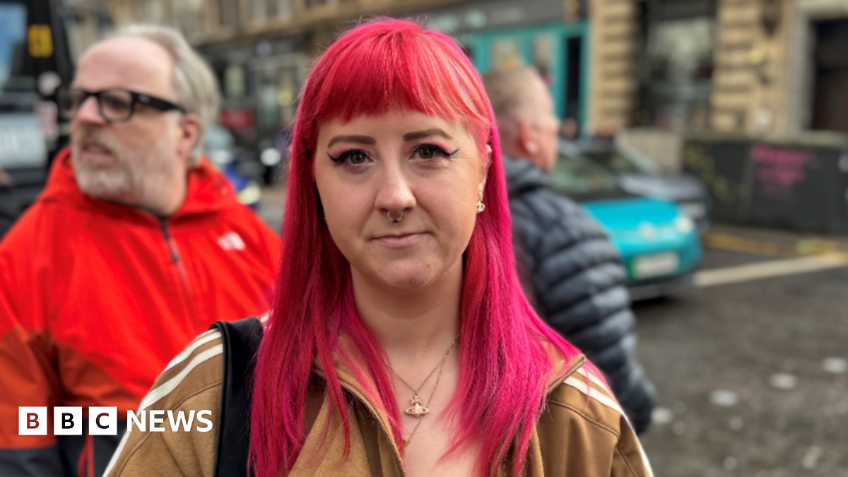 A woman with pink hair stands in the middle of the street with a light brown Adidas coat on