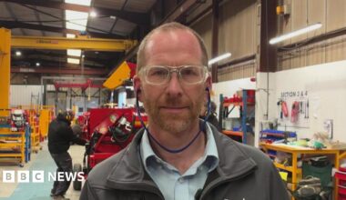 A man stands in a factory assembly area , behind him a man puts the final touches to a large piece of farm machinery. Tom wears protective glasses and ear plugs