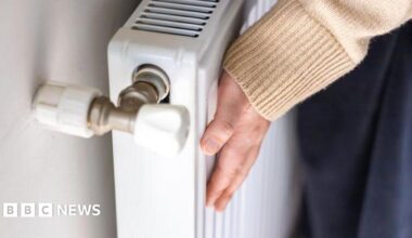 A woman touches a radiator in a house to check how warm it is. She is wearing a beige jumper. The close-up picture shows her hand and sleeve.