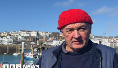 Richard Keen looking at the camera at the Fish Quay with small fishing boats behind him. He is wearing a navy jumper, a blue gillet and a red woolly hat.