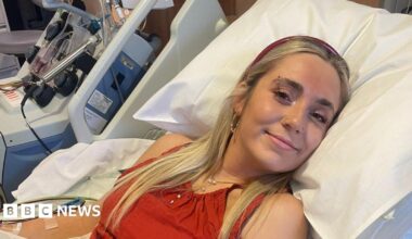Beth Bowyer is laying down in a hospital bed and is wearing a red top and headband. She's smiling at the camera.